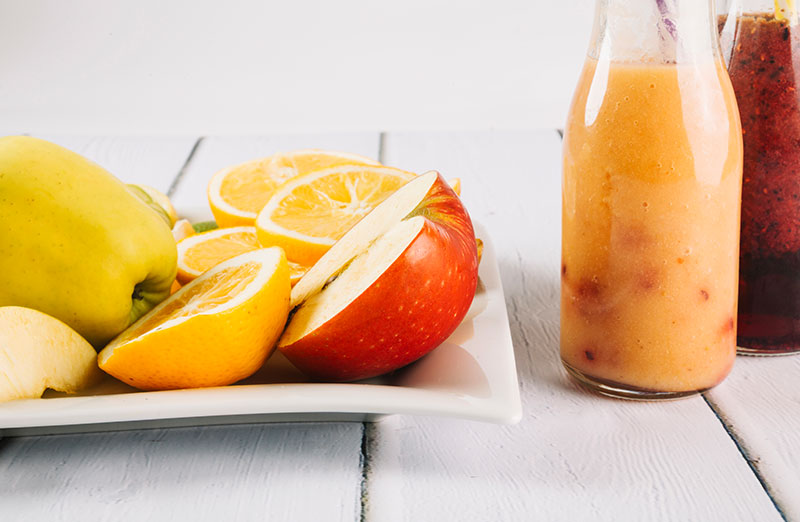 Botellas de cristal con zumo de frutas esterilizado, junto con un plato con manzanas y naranjas troceadas.